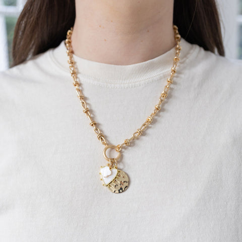 Model wearing a gold toggle chain with a large round textured pendant and two smaller charms, one a white heart and another a small white four leafed clover.