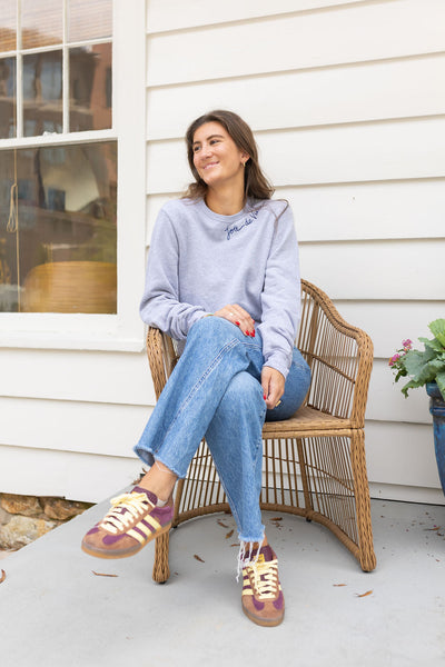 Woman sitting in a chair on a porch wearing a gray sweatshirt with blue stitching that says  "Joie De Vivre"