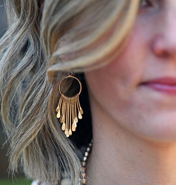 Picture of model wearing a gold hoop earring with gold bars in various lengths hanging from it. It gives the appearance of a sun catcher.