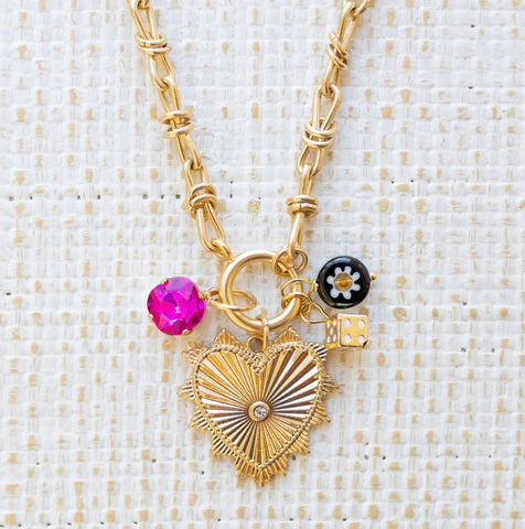 Picture of gold toggle chain with gold heart pendant with diamond in the center. Three other charms are attached, one a pink cut bead, a black bead with a white flower and a gold dice.