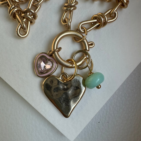 Picture of gold chain link charm necklace.  Necklace has a large gold heart pendant. Two smaller charms include a muted turquoise bead and a diamond heart charm with a pink and gold border.