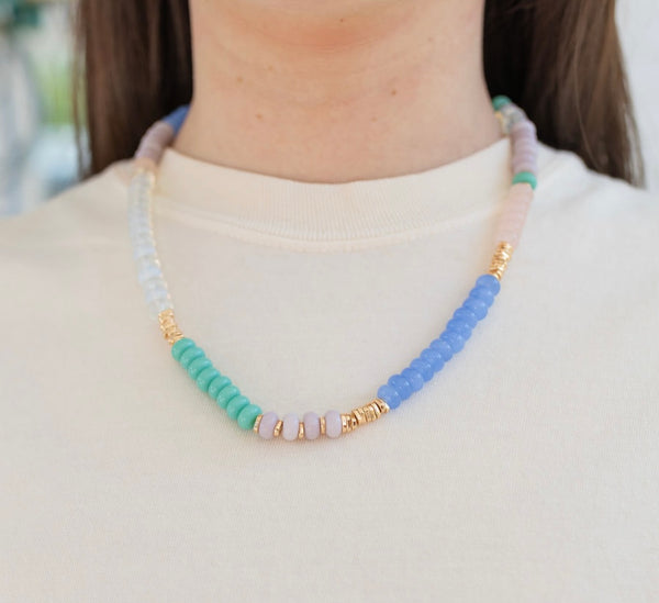 Model wearing a beaded necklace accented with small gold beads. Beads are white, rose pink, turquoise and blue.