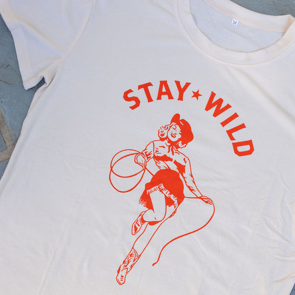 A cream colored t-shirt with a graphic of an orange cowgirl with a cowboy hat and lasso with the text "stay wild"