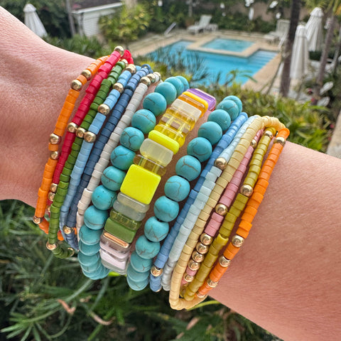 Picture of arm with a bundle of twelve colorful small beaded bracelets, two large bead turquoise bracelets and one square beaded bracelet