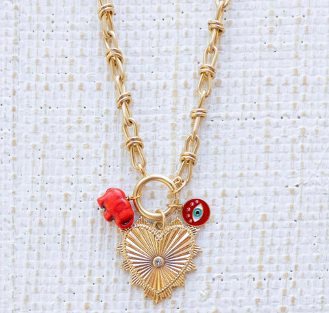 Picture of gold toggle chain with a large heart pendant with a diamond in the center and two charms. Charms are a red elephant and a red evil eye.