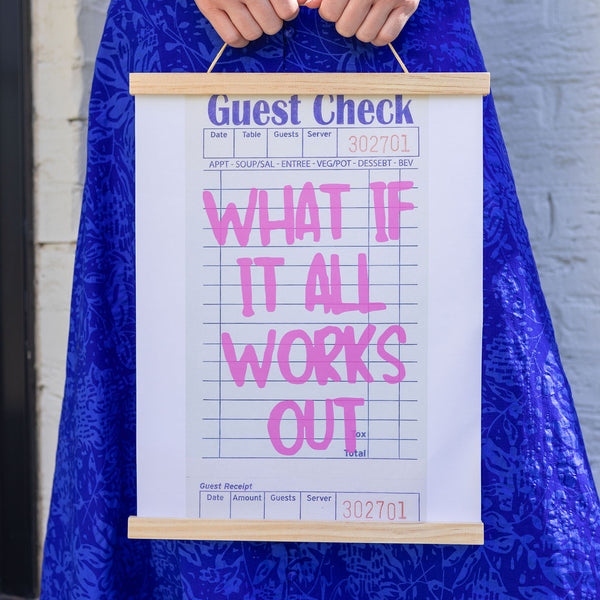 Girl against a wall wearing a blue dress holding a canvas hanging art piece that mimics a guest check with the text "what if it all works out" on a canvas 