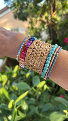 Picture of arm with 5 bracelets and one tan, woven cuff. Beaded bracelets are turquoise, coral, multicolored, blue and orange.