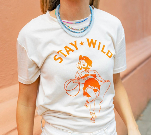Woman wearing a cream shirt with an orange print of a cowgirl with a lasso and the text "Stay Wild" with multiple blue beaded necklaces