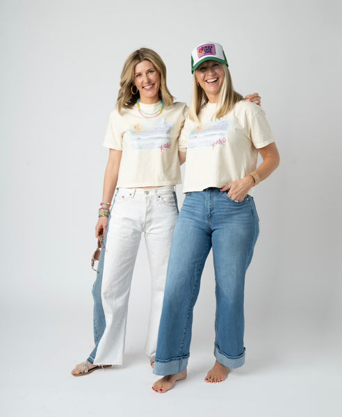 Picture of two women wearing jeans and matching beige cropped t shirts that have a beach scene.