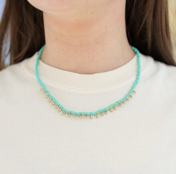 Model wearing a short necklace made of small turquoise beads and gold tear drop accent beads.