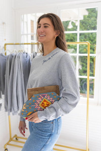 Woman standing in a house posing wearing a gray sweatshirt with blue stitching that says  "Joie De Vivre"