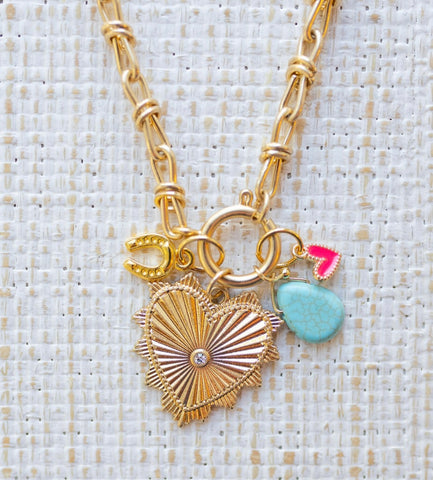 Picture of gold toggle chain with a large gold heart pendant with a diamond at center and several charms. Charms include a gold horseshoe, turquoise tear drop and small pink heart.