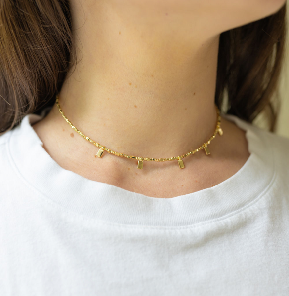 Girl wearing a gold necklace with a thin chain and little gold rectangles attached to it around her neck