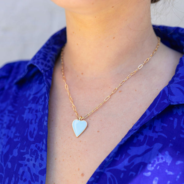 Woman wearing a thin gold chain necklace with a silver/light blue stone in the shape of a heart attached to it