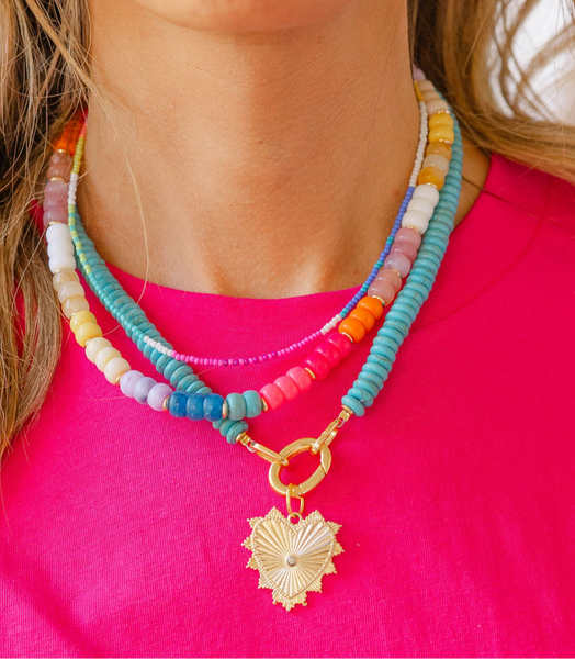 Girl wearing three necklaces: a big blue beaded necklace with a gold heart shape medallion attached to it, a multicolored beaded short necklace, and a thin multicolored beaded choker