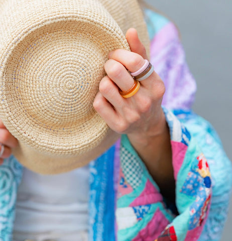 Woman wearing a colorful quilted robe holding a straw sun hat in her arms against her chest and wearing orange, blue, and pink rings 