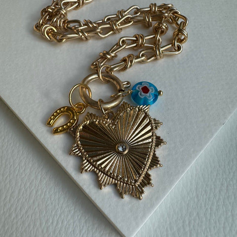 Picture of chunky gold chain necklace with a large gold heart pendant with a diamond stud in the center. Necklace also has two other charms attached, one a golden horseshoe and the other a blue bead with a flower design.